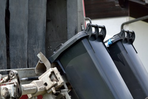 The image shows two black plastic wheelie bins positioned outdoors, attached to a metallic waste collection vehicle. The bins are made of smooth, durable plastic with a matte finish, featuring rounded corners and hinged lids, one of which is slightly open. The vehicle's metallic frame, visible on the left, has a silvery, slightly weathered surface with mechanical components and hinges connecting the bins to the loading mechanism. These bins are placed on a paved or concrete surface, likely at a commercial waste removal site in Tufnell Park, suggesting readiness for collection or disposal, with a nearby wooden structure partly visible in the background. The environment appears well-lit, possibly by natural daylight, casting mild shadows on the scene, which aligns with the professional service provided by Commercial Waste Tufnell Park for rubbish removal services in the area.