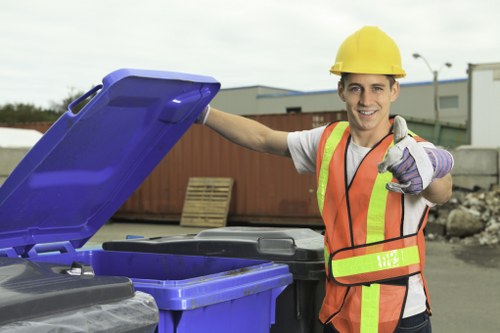 A young male waste management worker wearing a yellow safety helmet, high-visibility orange vest with reflective strips, and purple work gloves, is standing outdoors next to large blue and black rubbish bins on a paved area. He is smiling and pointing towards the camera with his right hand, while his left hand rests on the open lid of a blue bin. In the background, there are industrial buildings, wooden pallets, and a cloudy sky, indicating an urban or commercial environment. The scene suggests a rubbish collection or waste disposal operation, with the worker involved in waste management services typical of commercial waste removal in Tufnell Park or similar North London locations. The image is well-lit with natural daylight, highlighting the textures and colors of the equipment, clothing, and surrounding environment, supporting the context of professional waste collection services provided by Commercial Waste Tufnell Park.