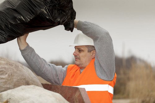 A waste management worker dressed in a grey long-sleeve shirt and an orange high-visibility vest, wearing a white safety helmet, is shown lifting and loading a black rubbish bag onto a pile of mixed waste materials outdoors. The waste pile in front of him comprises various items, including brown and beige bags, cardboard, and plastic, set on a rough, rocky surface. The background features an overcast sky and a blurred outdoor landscape, suggesting a rural or semi-urban environment. The scene aligns with rubbish removal services provided by Commercial Waste Tufnell Park, focusing on waste collection and disposal in the area around N7 postcode, near Tufnell Park. The worker’s task appears to involve the collection of household or general waste from an outdoor setting, indicative of professional rubbish removal operations in North London, supporting local waste management needs.