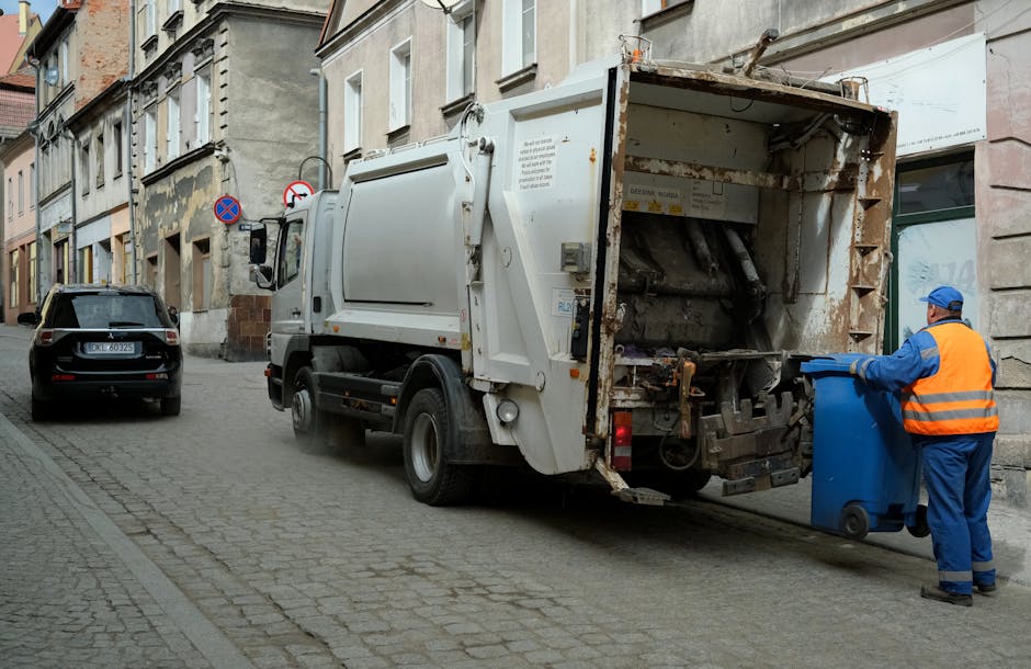 A large collection of mixed waste, including cardboard boxes, paper bags, plastic bags, and various discarded packaging materials, piled haphazardly around a communal rubbish bin on a paved urban stre