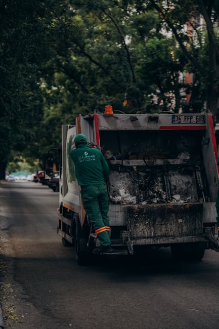 A waste collection vehicle parked on a suburban road with a worker in green uniform and safety helmet accessing the rear cargo area, which is open and filled with black, sooty debris, possibly indicat