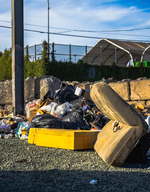 A middle-aged man with dark hair, wearing a short-sleeved black T-shirt with white text on the front, is seen on a paved sidewalk, bending over to dispose of waste into a large, cylindrical, stainless