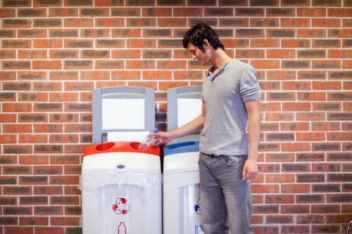 A young man with dark hair, wearing a light grey long-sleeved shirt and grey jeans, standing outdoors on a paved surface in front of a red brick wall, is disposing of a clear plastic bottle into a recycling bin. To his left are two recycling containers placed side by side; the first is white with a red recycling symbol and a circular opening, while the second is predominantly white with a blue recycling symbol and a matching opening. The environment appears to be in an urban or residential area, and the scene emphasizes responsible waste disposal, relevant to commercial waste management services provided by Commercial Waste Tufnell Park, serving the local postcode area around Tufnell Park and surrounding districts.