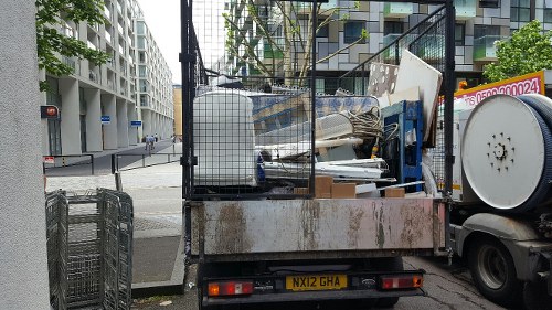 A flatbed truck parked on a city street in Tufnell Park, loaded with various types of waste materials. The load includes large, white, rectangular panels, possibly made of wood or plastic, alongside a silver metal box and scattered debris. The truck has a rusty flatbed with protective metal fencing at the sides and rear to secure the rubbish. Nearby, there are industrial metal waste cages and a street corner with modern, multi-storey residential buildings visible in the background, along with a traffic light and a pedestrian crossing. The scene is lit by natural daylight, indicating typical urban rubbish collection activity in North London.